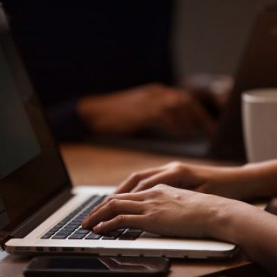 A photo of hands typing on the keyboard of a laptop, with a coffee cup in the background.